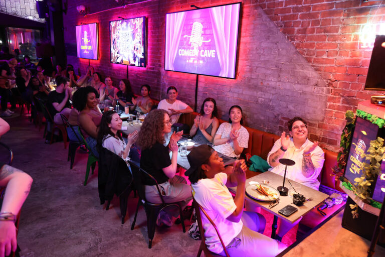 Audience enjoying Comedy Cave night at Teddy Beer with lively performances and bright neon lights.